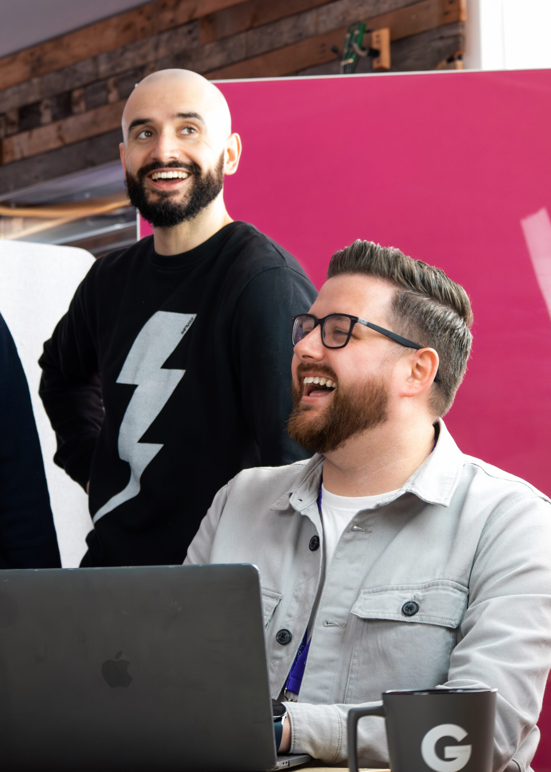 two men smiling by a laptop one sitting down and the other stood behind him